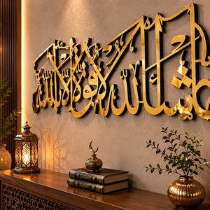 May include: A gold-colored Arabic calligraphy wall art piece with a black outline, mounted on a beige wall. The artwork is above a wooden console table with decorative items, including a lantern, books, and a small vase with greenery. The room has a modern, elegant aesthetic.