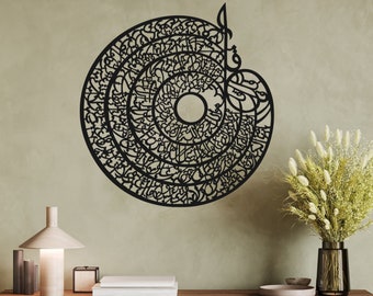 May include: Black metal wall art featuring a circular design with intricate Arabic calligraphy. The design is reminiscent of a mandala or a sunburst. The art piece is displayed on a beige wall with a wooden table and a vase of dried flowers in the foreground.