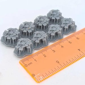 May include: A set of eight small, gray, 3D-printed miniature structures, each featuring a cluster of flat, square-shaped roof tiles supported by thin pillars on a hexagonal base. An orange plastic ruler shows the scale in centimeters.