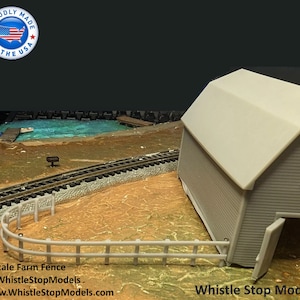 May include: N Scale model farm fence made of gray plastic. The fence is curved and has a series of vertical posts with horizontal rails. The fence is in front of a gray barn with a white roof. The text "Whistle Stop Models" is visible in the bottom right corner of the image.