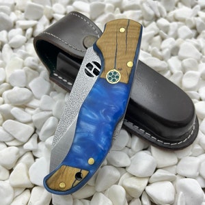 Könnte beinhalten: Ein blaues und braunes Taschenmesser mit einem Holzgriff und einer Lederscheide. Das Messer hat eine strukturierte Klinge und ein dekoratives Design am Griff.