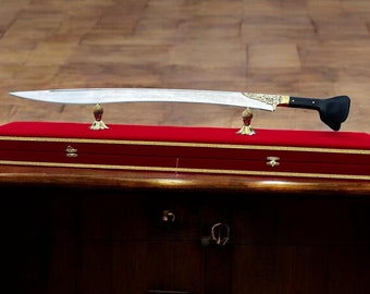 May include: A silver sword with a black handle and gold accents is displayed on a red velvet box with gold trim. The sword is resting on two gold metal stands.