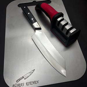 May include: A silver cutting board with a black and red knife sharpener and a black handled knife with a silver blade. The cutting board has the text "Büsra's Kitchen" engraved on it.