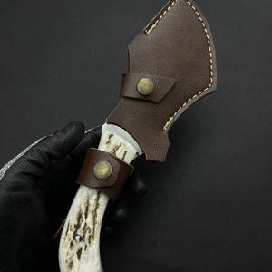 May include: A brown leather sheath with a white bone handle knife. The knife has a metal blade and a brass button on the sheath.
