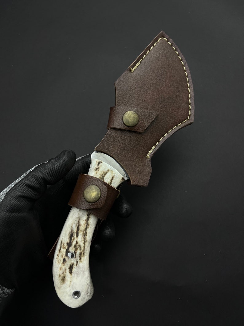 May include: A brown leather sheath with a white bone handle knife. The knife has a metal blade and a brass button on the sheath.