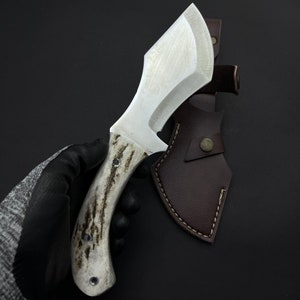 May include: A white-handled knife with a brown leather sheath. The knife has a curved blade and a textured handle.