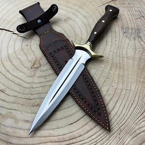 Puede incluir: Una daga con una hoja de acero pulido y un mango de madera oscura con detalles de latón. La daga está en una funda de cuero marrón con costuras decorativas y una trabilla para el cinturón. La hoja mide unos 25 cm.