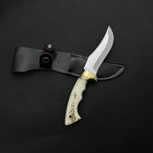 Peut inclure: Un couteau de chasse avec un manche blanc et brun et un fourreau en cuir.