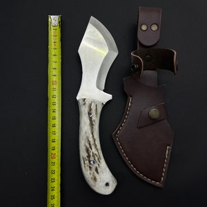 May include: A white-handled knife with a brown leather sheath. The knife has a silver blade and a white handle with a brown pattern. The sheath has a brown leather strap with a metal buckle.