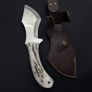 May include: A white-handled knife with a brown leather sheath. The knife has a curved blade and a white handle with a dark brown pattern. The sheath has a brown leather strap with two brass snaps.