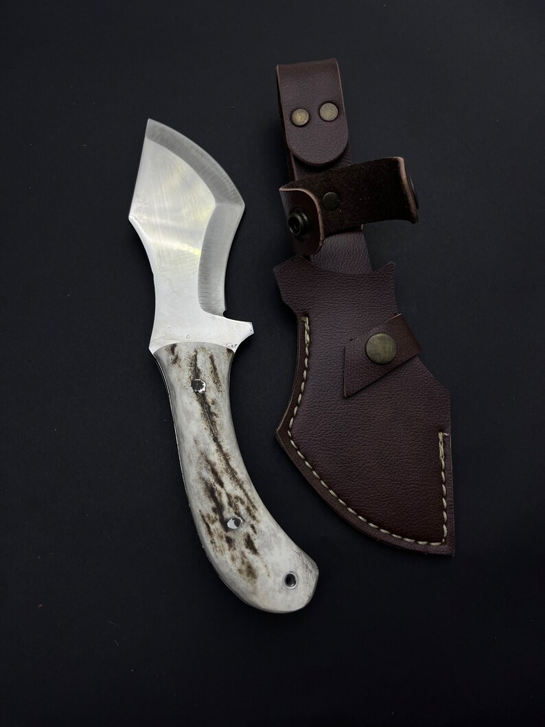 May include: A white-handled knife with a brown leather sheath. The knife has a curved blade and a white handle with a dark brown pattern. The sheath has a brown leather strap with two brass snaps.