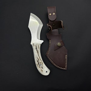 May include: A white-handled knife with a brown leather sheath. The knife has a curved blade and a white handle with a dark brown pattern. The sheath has a brown leather strap with a metal buckle.