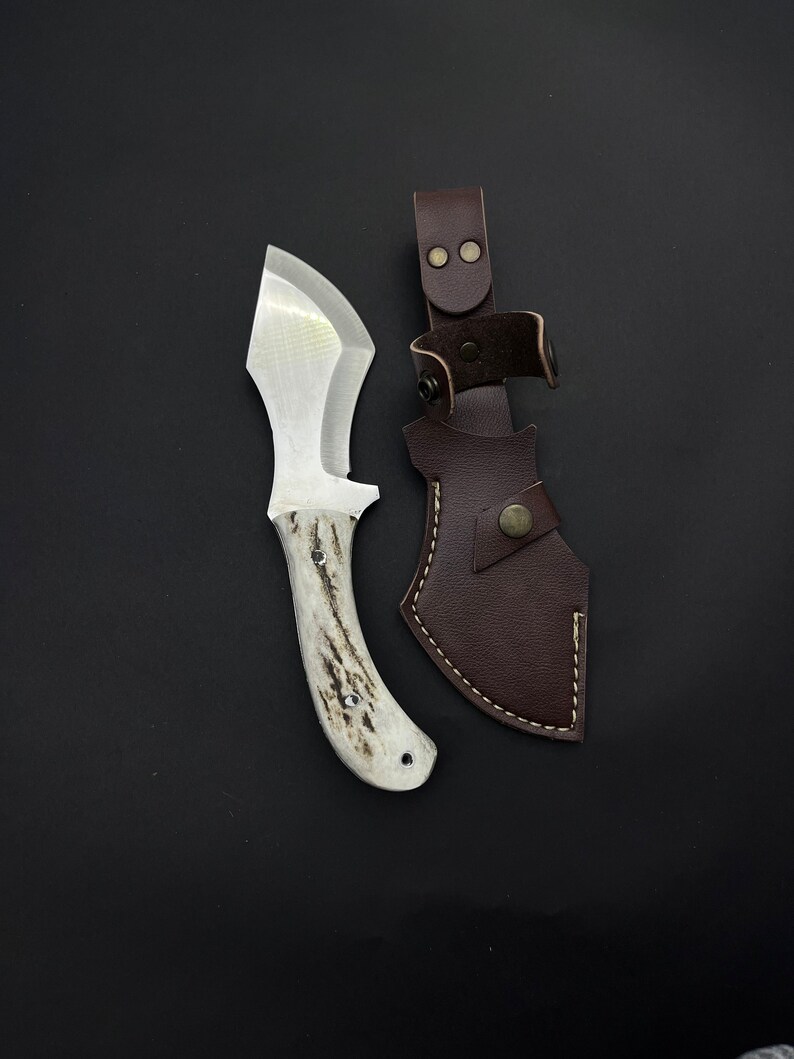May include: A white-handled knife with a brown leather sheath. The knife has a curved blade and a white handle with a dark brown pattern. The sheath has a brown leather strap with a metal buckle.