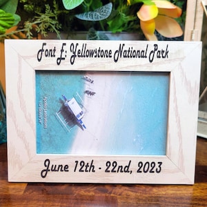 May include: A light-colored wooden picture frame with a visible grain pattern. The top border is engraved with the text Font E: Yellowstone National Park, and the bottom border features the engraved text June 12th - 22nd, 2023. The frame holds a photo of a boat.
