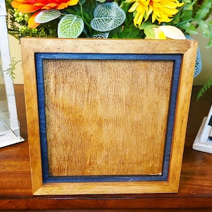 May include: Square wooden picture frame with a natural wood finish and a dark blue inner border. The frame is empty, ready for a photo or artwork. The frame is sitting on a wooden surface.