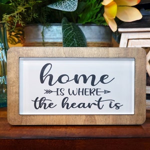 May include: A wooden sign with a white background and black lettering that reads "home is where the heart is". The sign is framed in a light brown wood frame.