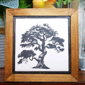 Può includere: Un oggetto da parete quadrato con una piastrella in ceramica bianca che presenta un'illustrazione a inchiostro nero di un bonsai nodoso. La piastrella è inserita in una cornice interna scura e una cornice esterna in legno chiaro con venature.