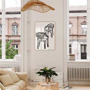 May include: A framed art print featuring a minimalist black and white illustration of two chairs. The artwork is displayed in a bright, airy living room with a beige sofa, wooden coffee table, and a woven light fixture.