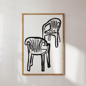 May include: Black and white art print featuring two stylized chairs. The chairs are depicted with bold, brushstroke-like lines, creating a minimalist and modern aesthetic. The artwork is framed in a natural wood frame, adding a touch of warmth.