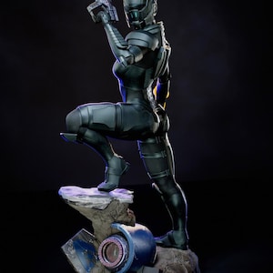 Puede incluir: Estatua detallada de una figura femenina con armadura futurista, sosteniendo un arma. La figura posa sobre una base rocosa con un fondo oscuro. La armadura es principalmente negra con detalles en rojo y azul. El logotipo de KC Studio es visible.