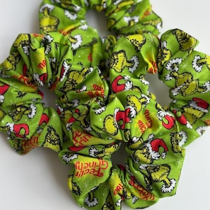 May include: Four green scrunchies with a repeating pattern of a cartoon character wearing a Santa hat. The text "Feel Grinchy" is visible on the fabric. The scrunchies are made of a soft, gathered fabric.