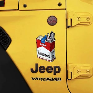 May include: A yellow Jeep Wrangler Unlimited vehicle panel featuring a Trail Rated 4x4 badge and a sticker depicting a red and white pack of cigarettes labeled Motorsport, with spark plugs protruding from the top and a piece of blue tape.
