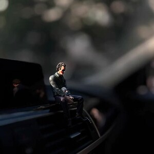 May include: A small figurine of a character with dark hair, wearing a dark green jacket and dark trousers, sitting on a black car air vent. The figure has a pale face with red makeup around the mouth and is holding a small white skull object.