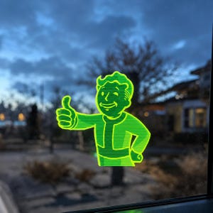 May include: A bright neon green vinyl decal of the Fallout Vault Boy character, featuring a winking expression and a thumbs-up gesture. The design is composed of horizontal lines, creating a striped effect on the character's figure.