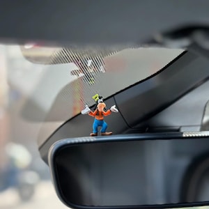 May include: A small Goofy figurine standing on top of a car rearview mirror. The character wears a bright orange sweater, blue pants, brown shoes, and white gloves, with a tall green hat perched on its head, arms outstretched in a welcoming pose.