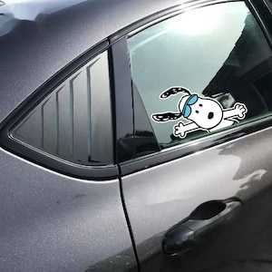 May include: A white vinyl decal of a cartoon dog with long black ears and blue goggles applied to the rear passenger side window of a dark gray car. The dog is depicted with arms outstretched as if pressing against the glass surface.