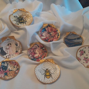 May include: Eight decorative seashells with gold trim, each featuring a different design. The designs include bees, flowers, and a woman in a dress.