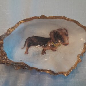 Puede incluir: Un plato de concha de ostra con borde dorado con una imagen pintada de un dachshund con un collar amarillo.