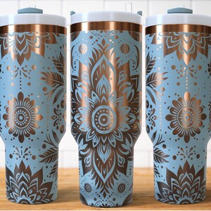 May include: Three light blue insulated tumblers with copper-coloured floral designs. Each tumbler has a white lid, a handle, and a copper-coloured band. The floral design includes flowers, leaves, and dots. The tumblers are on a wooden surface.