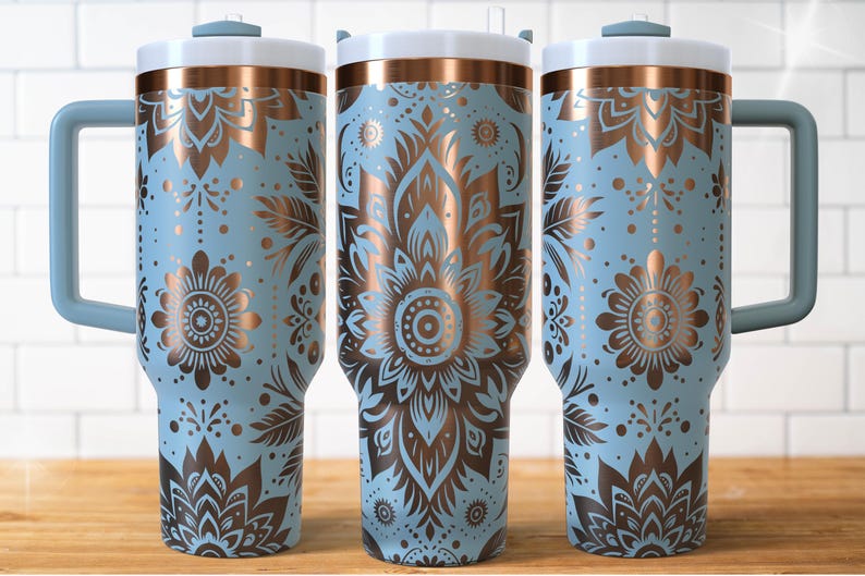 May include: Three light blue insulated tumblers with copper-coloured floral designs. Each tumbler has a white lid, a handle, and a copper-coloured band. The floral design includes flowers, leaves, and dots. The tumblers are on a wooden surface.