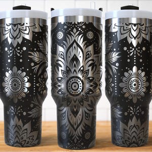 May include: Three black insulated travel mugs with silver floral patterns. Each mug has a white lid, a black handle, and a black rim. The floral design includes flowers, leaves, and dots. The mugs are on a wooden surface.