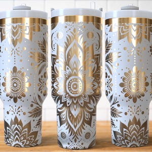 May include: Three white insulated tumblers with gold floral patterns and grey handles. The design features detailed gold flowers, leaves, and geometric shapes. Each tumbler has a white lid and a straw.