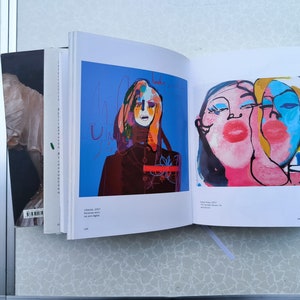 May include: Two pages from an art book featuring colourful abstract paintings of faces. The first page shows a woman with a red and black outfit against a blue background. The second page shows two faces with pink, blue and yellow paint.
