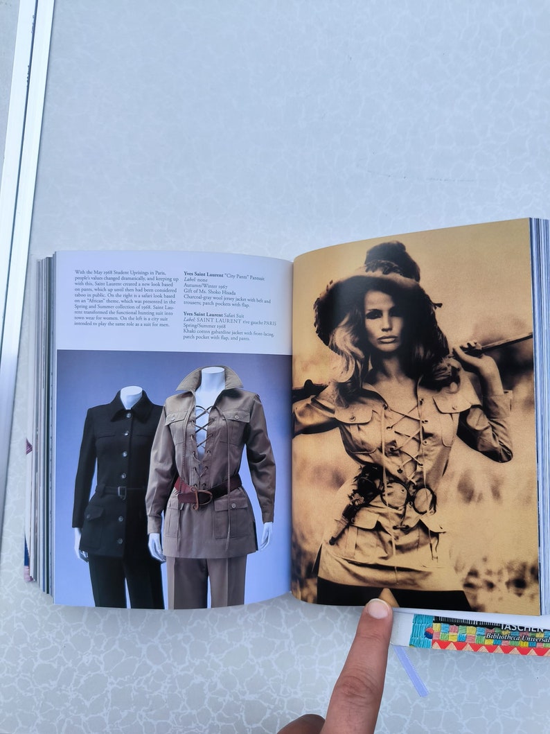 May include: Two models wearing safari-style jackets. The jacket on the left is black and has a button closure. The jacket on the right is brown with a lace-up front and a brown belt. The text "Yves Saint Laurent "City Farm"  Parfum" is visible in the image.