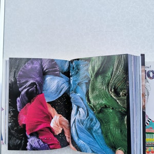 May include: An open book with colourful fabric pages. The fabric is folded and layered, creating a textured and abstract design. The colours include purple, pink, blue, and green.