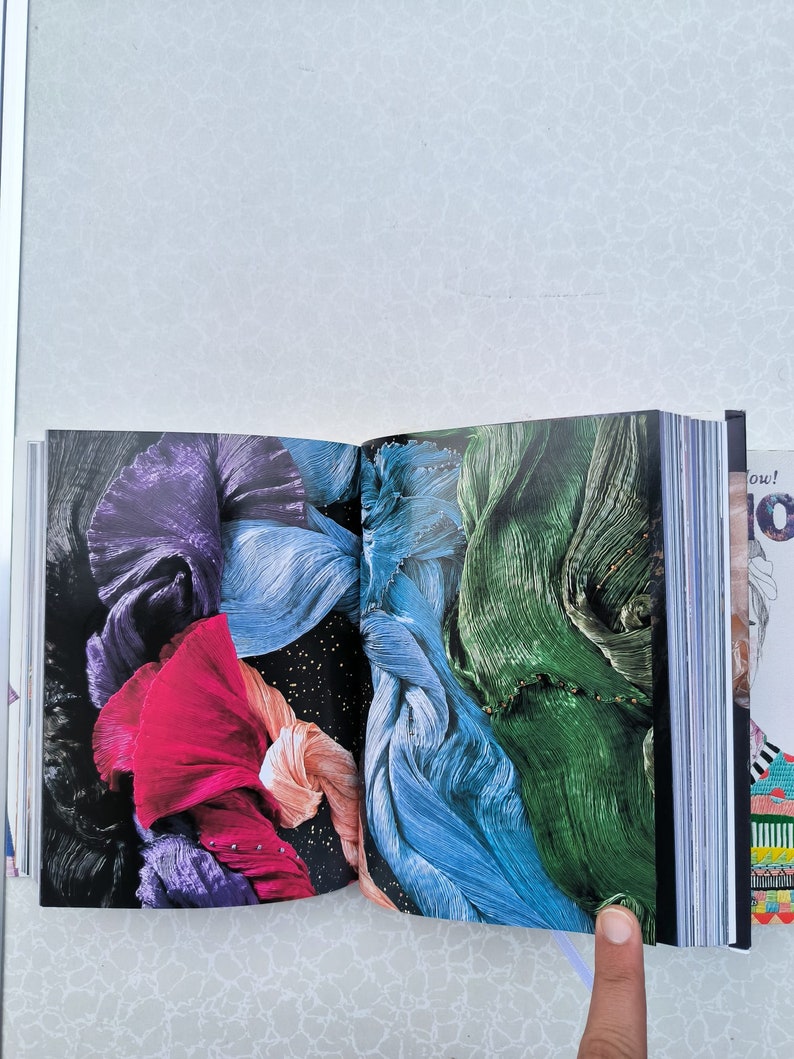 May include: An open book with colourful fabric pages. The fabric is folded and layered, creating a textured and abstract design. The colours include purple, pink, blue, and green.