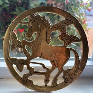 May include: A brass circular trivet with a cutout design of a unicorn standing on its hind legs. The unicorn is surrounded by stylized leaves.