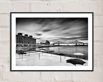 Puede incluir: Una fotografía en blanco y negro de una ciudad con un puente en la distancia. En primer plano, una playa con varias sombrillas negras.