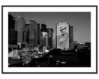Puede incluir: Fotografía en blanco y negro de un horizonte urbano con un gran mural de un hombre con sombrero en un edificio.