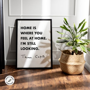May include: A rectangular framed wall art print with a white background and black text that reads: HOME IS WHERE YOU FEEL AT HOME. I'M STILL LOOKING. The signature Truman Capote is printed in black at the bottom of the white paper surface.
