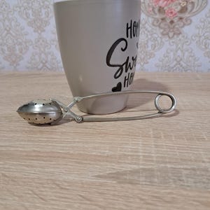 以下が含まれることがあります： 「Home Sweet Home」の黒い筆記体文字が入った灰色のセラミックマグカップと、銀色の茶こし。茶こしは、穴の開いた楕円形のバスケットと長いハンドルが付いています。