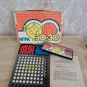 Puede incluir: Un juego de mesa vintage con una caja con personajes de dibujos animados y el texto "30-30". El juego incluye un tablero de plástico negro con clavijas, canicas rojas y azules e instrucciones. La caja es naranja y blanca.