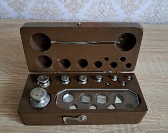 Puede incluir: Una caja de madera marrón antigua abierta que contiene varios pesos de metal. La caja tiene una tapa con bisagras y un compartimento con varios pesos circulares y rectangulares. Los pesos son de diferentes tamaños y formas, y algunos tienen marcas que indican su peso.
