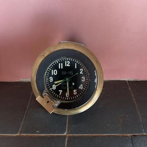 May include: A vintage clock with a black face, white numerals, and a brass-coloured bezel. The clock has a small, rectangular brass-coloured latch on the bottom left. The hands are white and green. The clock is round and sits on a dark surface.