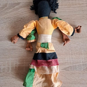 May include: Handmade cloth doll with a black head and curly hair. The doll wears a yellow top with green and black accents, a white belt, and a layered skirt in black, red, and green. The doll's arms and feet are made of a brown material.
