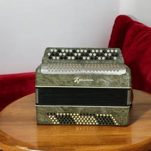 May include: An olive green accordion with black and white buttons and a silver accent. The accordion is resting on a round wooden table. The text "Крещение" is visible on the front.